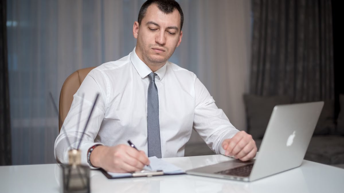 Professional businessperson in corporate attire writing at a white office desk with a laptop.